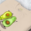 Close-up detail of a folded cream white Avocado Yoga Tee, highlighting the playful yoga‑avocado illustration with smooth lines and soft color tones.
