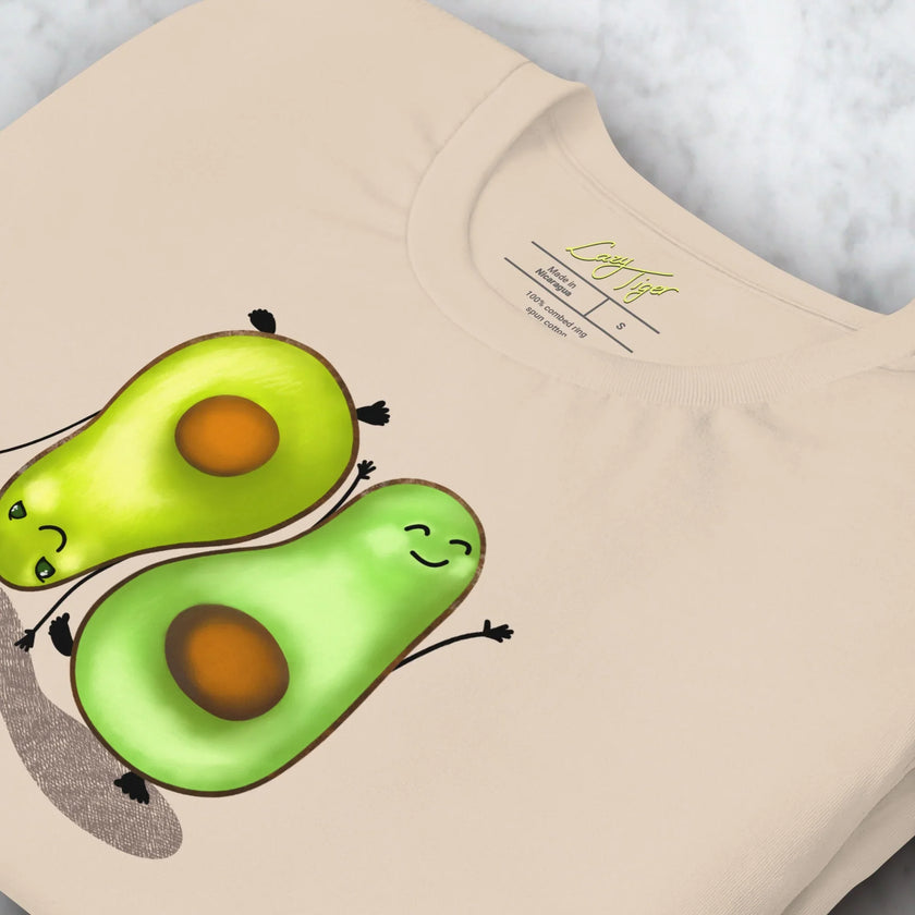 Close-up detail of a folded cream white Avocado Yoga Tee, highlighting the playful yoga‑avocado illustration with smooth lines and soft color tones.
