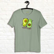 Sage green Avocado Yoga Tee hanging on a wall, showing two cartoon avocados doing yoga exercises with charming expressions and soft, aesthetic colors.