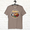 Taupe Lazy Potato Tee hanging on a wall, showing the relaxed potato on a red couch with the neon‑style ‘Couch Potato’ script above.