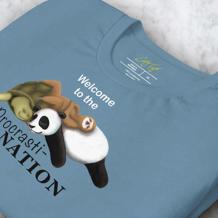 Detailed view of folded denim blue tee showing the Procrasti‑NATION sloth, panda, and turtle illustration.